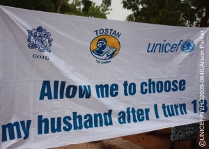 A banner bearing the slogan Allow me to choose my husband after I turn 18, and the logos of the Government, the NGO Tostan and UNICEF, advocates against early marriage. It is being displayed at an event in Darsilameh Village renouncing FGM/C and early marriage in 24 communities in eastern Upper River Region. The change in community practice of these traditional norms came about through their participation in the Community Development and Empowerment Programme, supported by the Government, the NGO Tostan and UNICEF.  On 14 June 2009 in the Gambia, women representing 24 neighbouring villages in Upper River Region gathered in Darsilameh Village to announce and celeate an end to the practices of female genital mutilation/cutting (FGM/C) and early marriage in their communities. An estimated 3 million African girls in 28 countries are subjected to FGM/C every year, a social convention linked to traditional perceptions of girls status for marriage. But FGM/C also causes great suffering and often life-long and life-threatening health risks. FGM/C and other pervasive practices such as early marriage are now recognized as manifestations of gender inequalities that threaten the well-being of girls and women and increase maternal health and child mortality risks. In the Gambia, 78 per cent of girls/women aged 15-49 years have been subjected to FGM/C; a figure that rises to 99 per cent in the Upper River Region. Likewise, almost half of Gambian girls marry before age 18. The 14 June celeation is part of a process, now underway in several African countries, of changing harmful and gender-discriminating social norms through a human rights-based approach, in which knowledge and support are offered to encourage positive change that is directed by community members themselves. In Upper River Region, some 80 communities  mainly from the Mandinka ethnic group  are participating in this process through the Community Development and Empowerment Programme, implemented by the Government, the international NGO Tostan and UNICEF. Public declarations renouncing harmful practices are a critical part of the process, affirming a communitys commitment and helping to create a critical mass for nationwide change. More than 600 people  including girls and women, religious leaders, village chiefs, delegates from youth and womens groups, government officials and representatives from Tostan and UNICEF  attended the Darsilameh celeation.