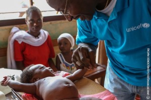 UNICEF Representative in the Central African Republic Souleymane Diabaté plays with a baby at the ADECOM (Association for Community Development) health post in the Bogbaya neighbourhood in Bangui, the capital, during the measles vaccination campaign. A woman and another child are seated nearby. Children are also receiving vitamin A and deworming tablets during the campaign. Mr. Diabatés shirt bears the UNICEF logo.  From 22 to 26 May 2013 in the Central African Republic, an emergency measles vaccination campaign is being held after eight children in Bangui, the capital, tested positive for the disease in April. The campaign is being implemented by the Ministry of Health, the World Health Organization (WHO), UNICEF and other partners and aims to immunize an estimated 125,000 children under age 5. Measles remains a leading cause of death among young children worldwide. In the Central African Republic, one of the worlds poorest countries, the measles vaccination rate stands at a low 62 per cent. Recent conflict has also contributed to a further eakdown in health and other services and put hundreds of thousands of children at risk of the disease. An estimated 246,500 vaccines have been despatched to Bangui in response to the outeak, and to improve routine measles vaccination in the countrys high-risk regions. In response to the current crisis, UNICEF is supporting emergency health activities in Banguis four main hospitals and at health centres throughout the country (including health supplies for up to 141,000 people for three months), as well as interventions in the areas of nutrition, water, sanitation and hygiene (WASH), education and child protection.