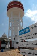A sign bearing the UNICEF logo stands near a newly built water tower in the southern village of Froun. The tower, which contains 300 cubic metres of water, was built with support from UNICEF and the NGO Islamic Relief Worldwide. UNICEF has helped repair and reconstruct water towers and reservoirs destroyed during last year's conflict, including restoring 37 water supply systems - benefiting almost 300,000 people - over the past year.  On 12 July 2007 in Lebanon, one year after the war between Israel and Hezbollah (a Lebanon-based political faction), renewed political instability and insecurity continues to impede children's recovery after the crisis. Thousands of children were affected by the 34-day conflict (from 12 July-14 August 2006), which killed an estimated 1,187 people, injured nearly 4,100 and left some 907,000 internally displaced. An additional 150,000 sought temporary refuge in neighbouring Syria. The conflict also significantly damaged infrastructure, including airports, roads, power plants, idges and fuel stations. Ongoing crises over the past several months include bombings throughout the country and conflict and displacement in Palestinian refugee camps in the north. Working with the Government, other UN agencies and NGOs, UNICEF continues to provide psychosocial support and basic counselling for traumatized children, and supports interventions in the areas of water, sanitation and hygiene, health, education and child protection. UNICEF assistance includes: training counsellors and teachers to identify traumatized children; providing essential learning materials to 400,000 students in 1,400 schools in support of the country's national Back-to-School campaign; establishing child-friendly spaces and securing alternative learning spaces for children as schools are rehabilitated; providing safe water and sanitation facilities for families and communities and promoting hygiene; repairing and reconstructing water supply systems; supporting immunization campaigns; assisting with shelter and other basic services, and supporting mine-risk education programmes and public mine-awareness campaigns warning children of the dangers of unexploded ordnance (UXOs). An estimated 1 million UXOs remain undetonated on roads and in homes and fields.